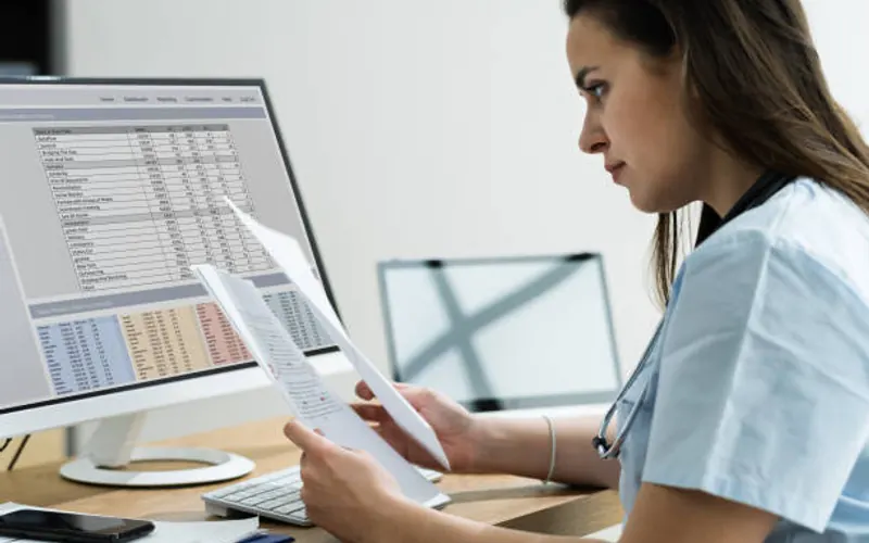 A person in scrubs checking a printed report against a record on screen